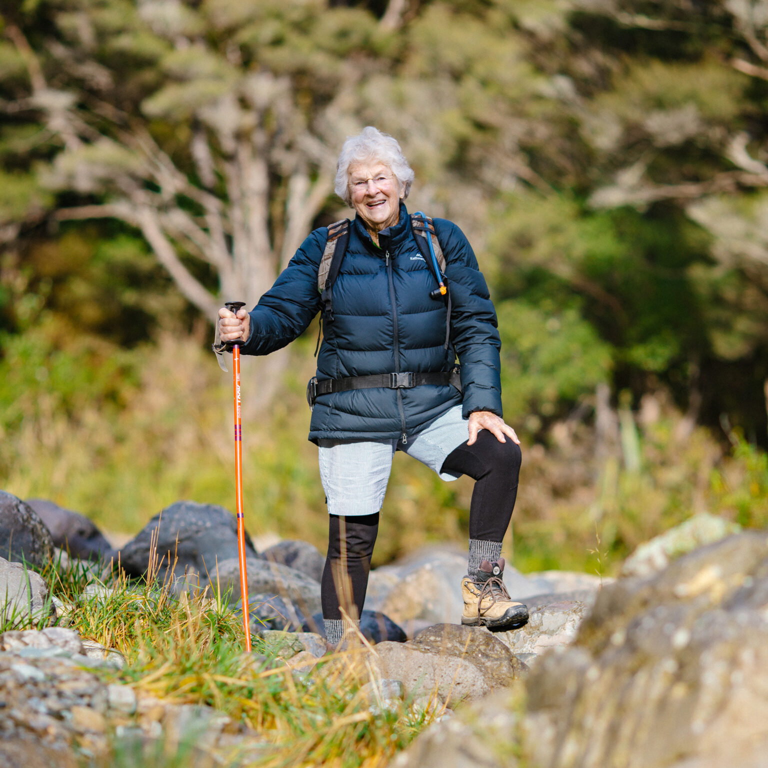 An elderly lady standing in tramping gear on a rock prouding